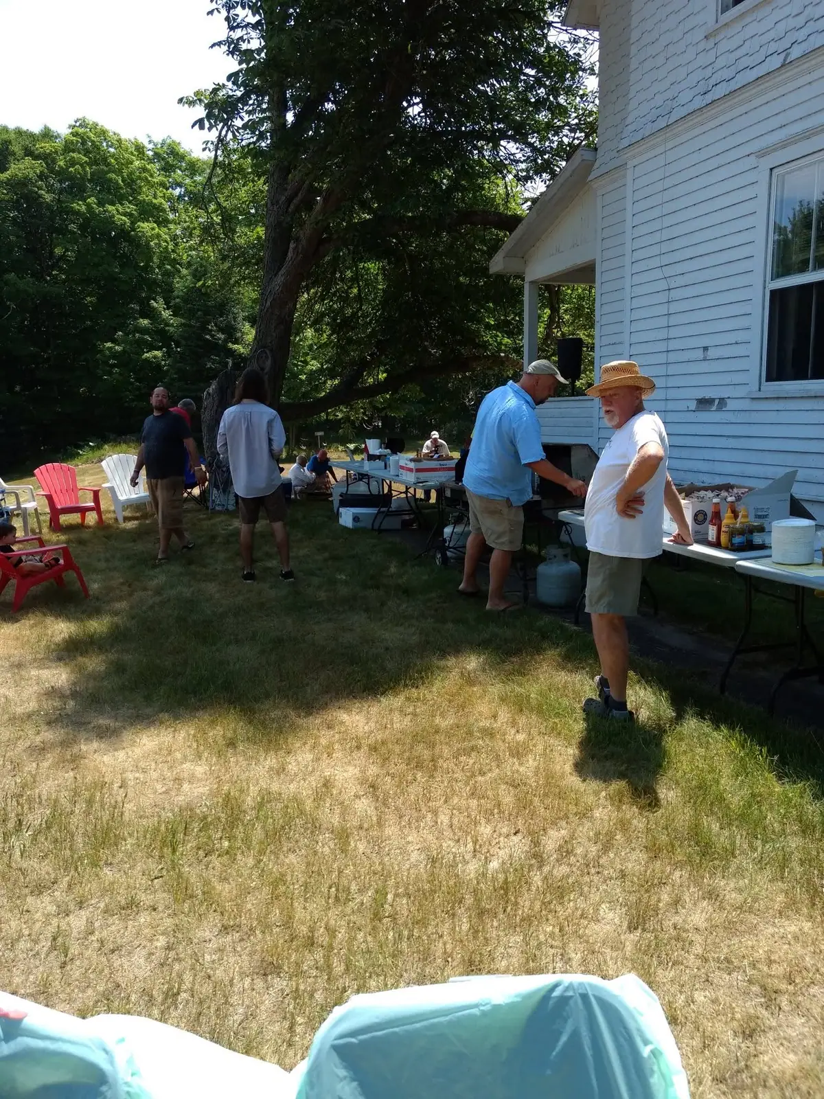 People gather beside a white clapboard house for an outdoor picnic with tables, chairs, and a grill.