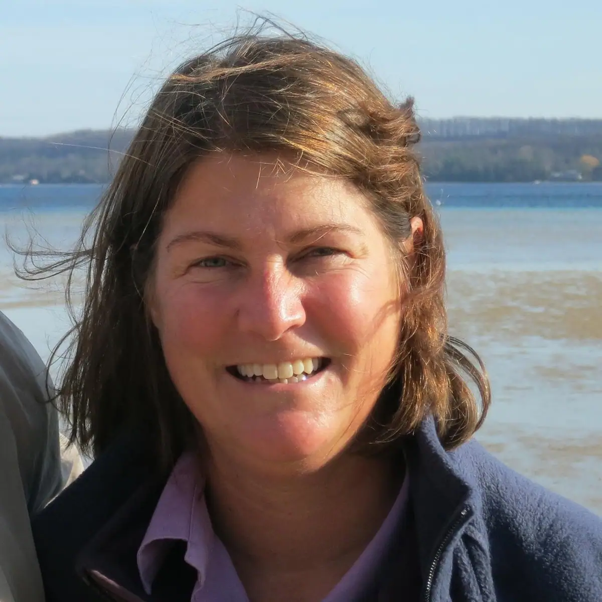 Smiling woman in a blue jacket photographed beside a broad body of water.