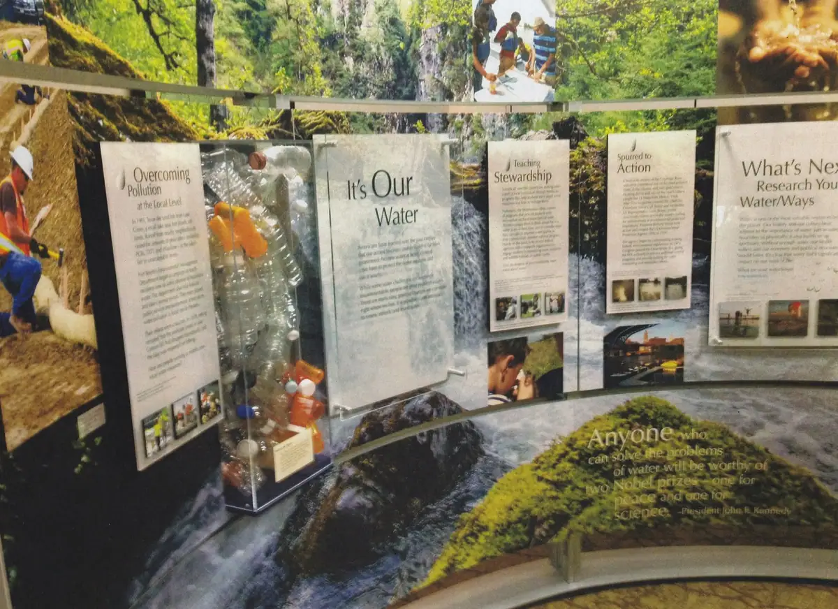 An indoor water-education exhibit with illustrated panels, a waterfall backdrop, and a case of plastic bottles.