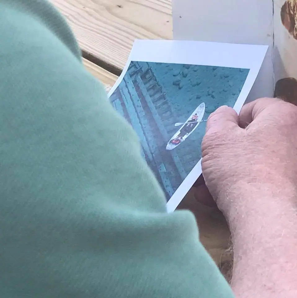 A printed photograph of a kayaker is held by two people over a wooden surface.