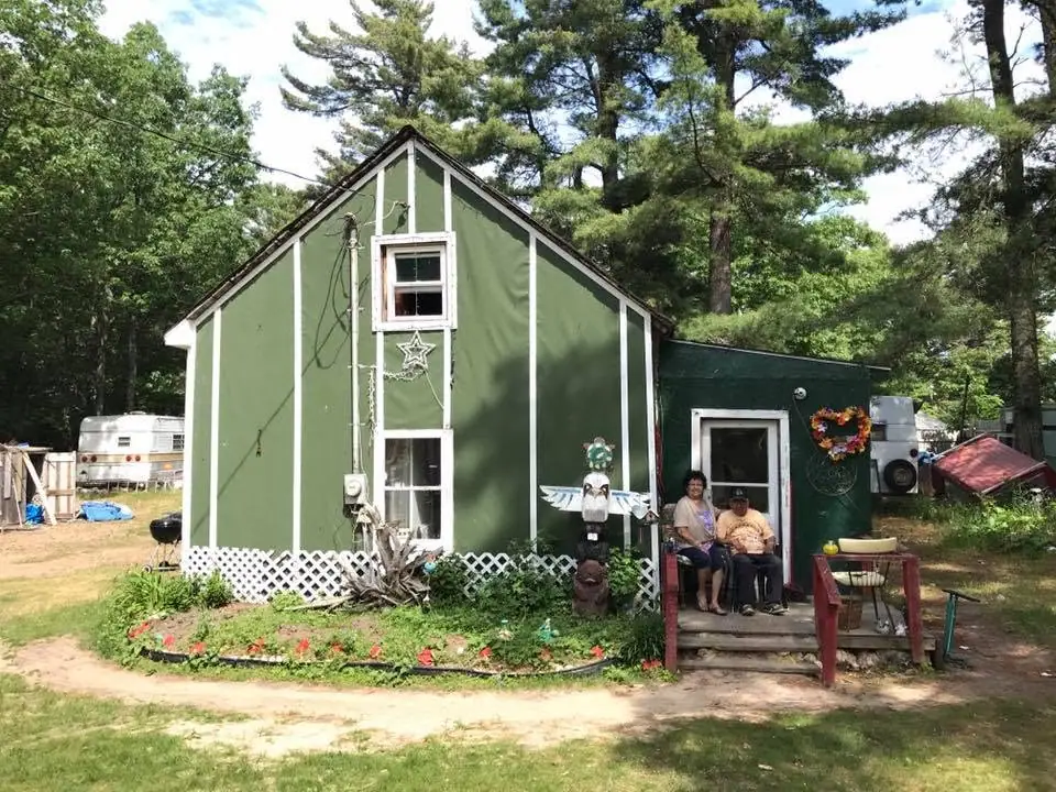 A green cabin with white trim, a flower garden, and two people seated on its porch.