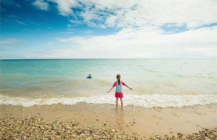 A child stands with arms outstretched at the edge of a pebbly beach as another person wades offshore.