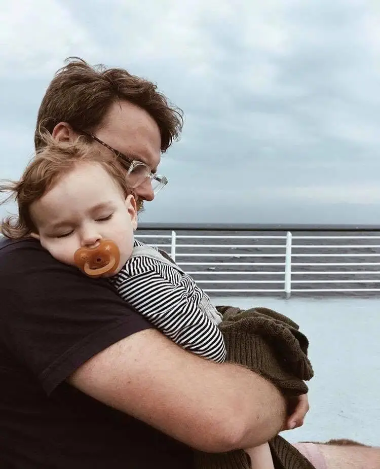 An adult holds a sleeping child on a boat deck beneath a cloudy sky.