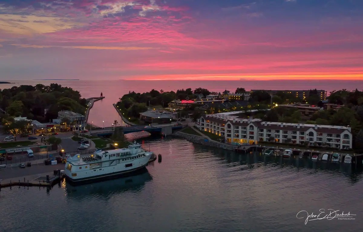 Aerial sunset view of a harbor with a ferry, docks, buildings, and a lighthouse.