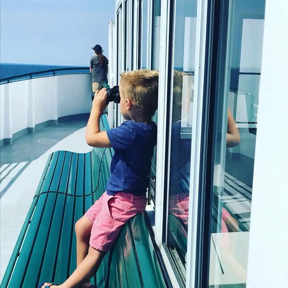 A child uses binoculars on a ferry deck while an adult stands at the railing overlooking open water.