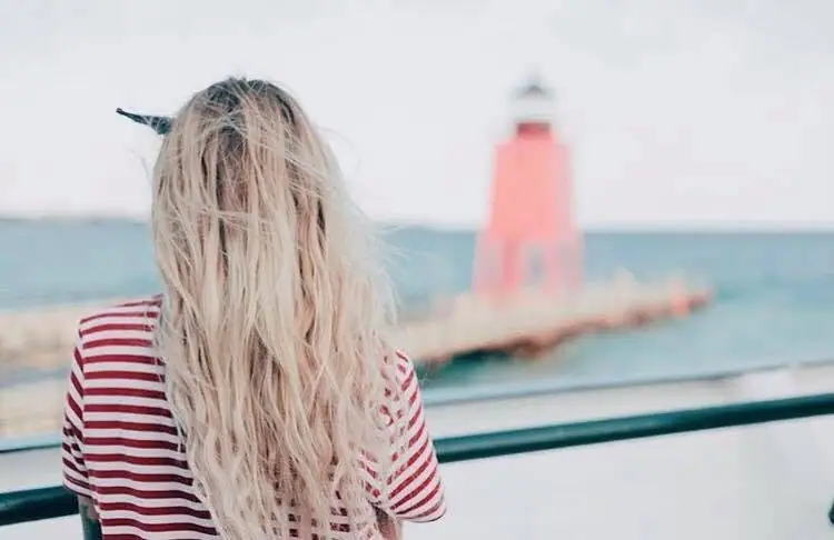 A person in a striped shirt looks toward a red lighthouse at the end of a pier.