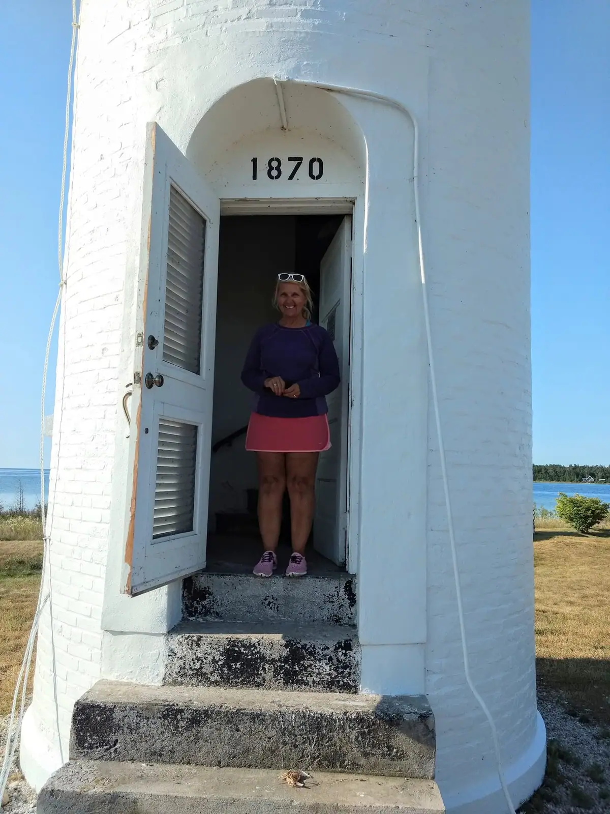 A smiling woman stands in the open doorway of a white brick lighthouse marked 1870.