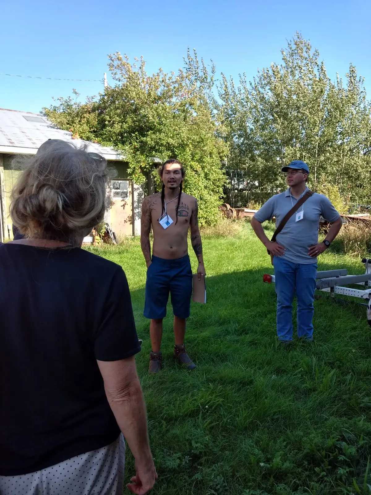Three adults gather outdoors beside a small building and utility trailer, with one holding a clipboard.