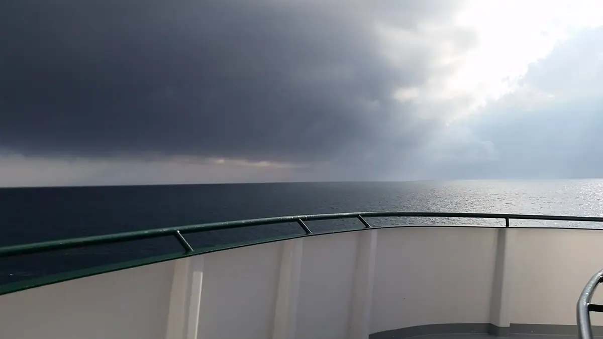 A ship’s curved deck railing overlooks dark open water beneath heavy storm clouds and a bright break in the sky.