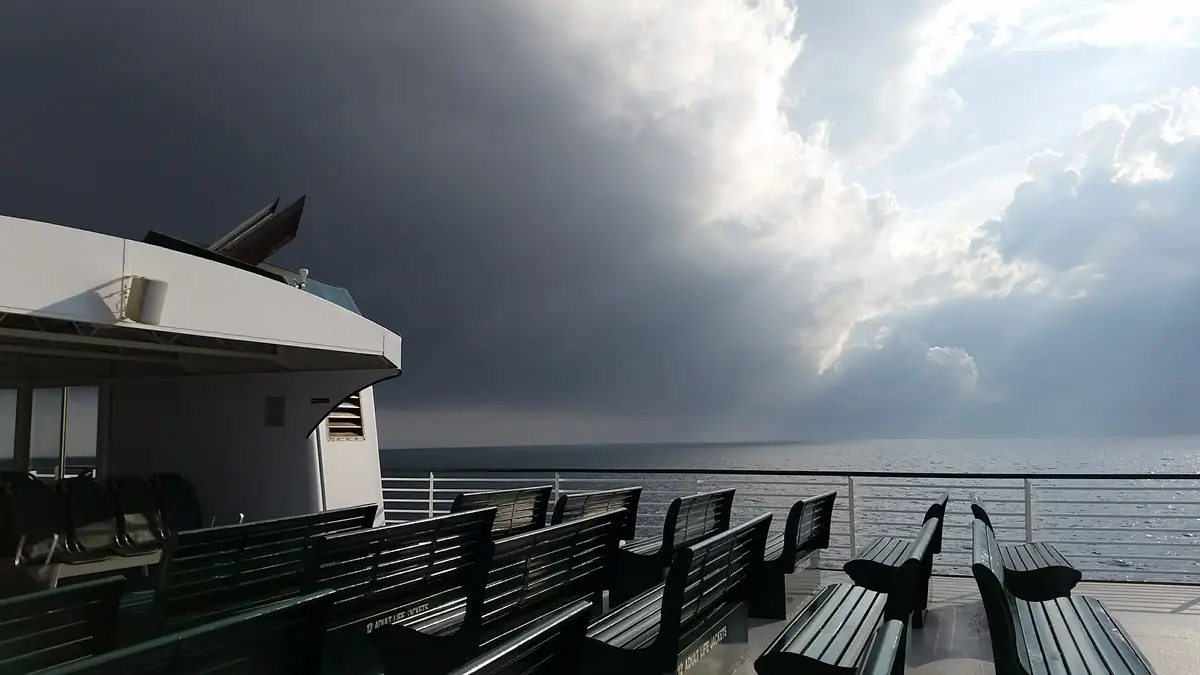 Green benches line a ferry deck beneath dark clouds and bright sunlight over open water.