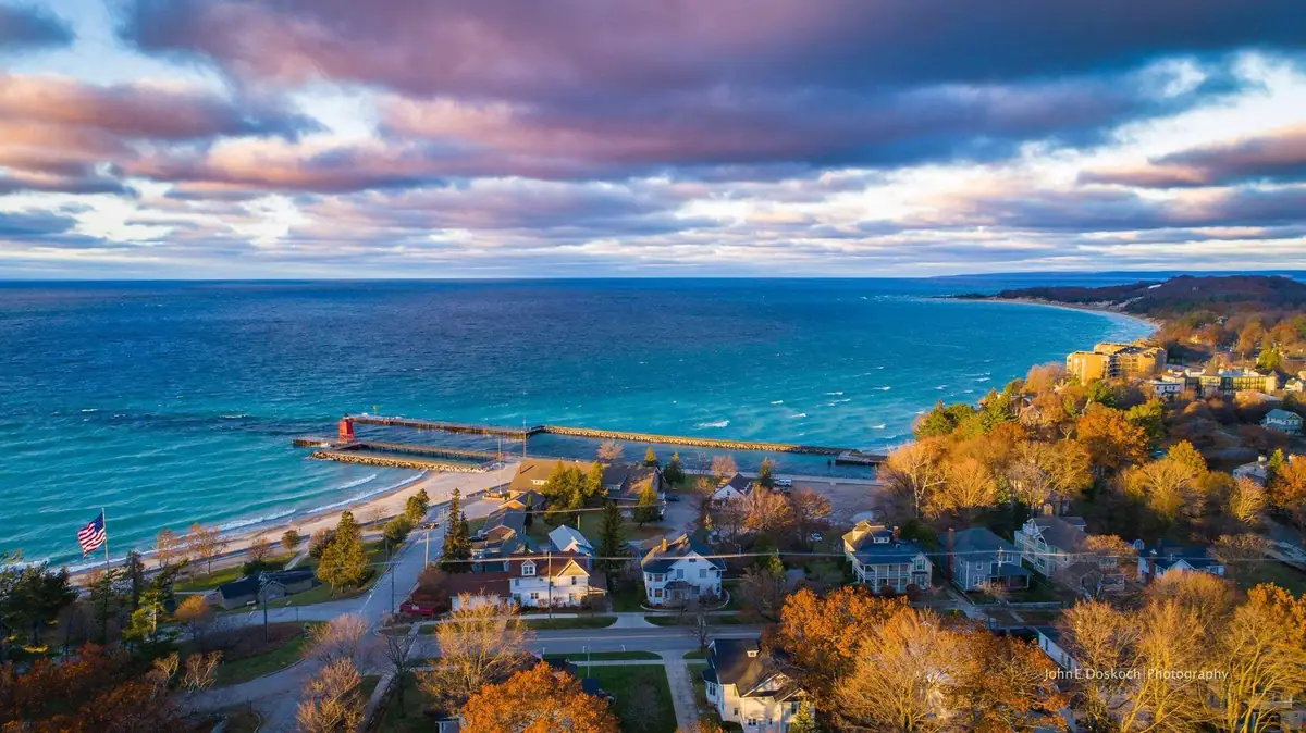 Aerial view of a waterfront neighborhood, pier, sandy beach, autumn trees, and dramatic clouds.