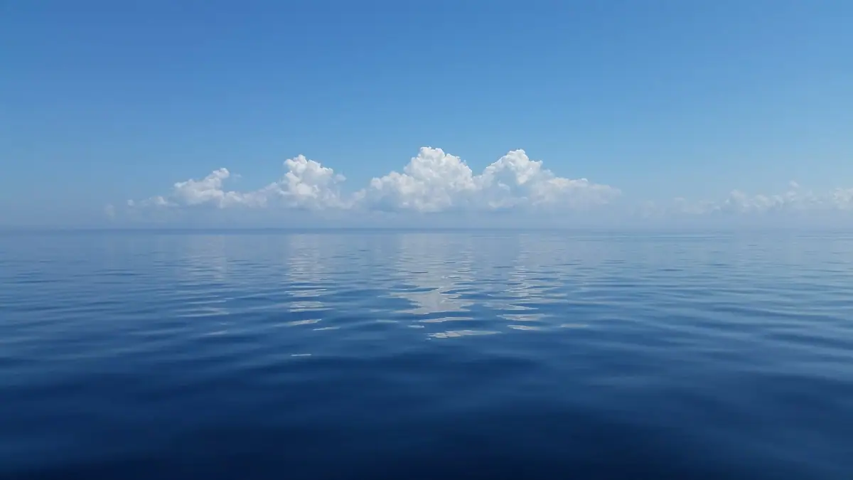 Calm blue water stretches to a low horizon beneath bright cumulus clouds and a clear blue sky.