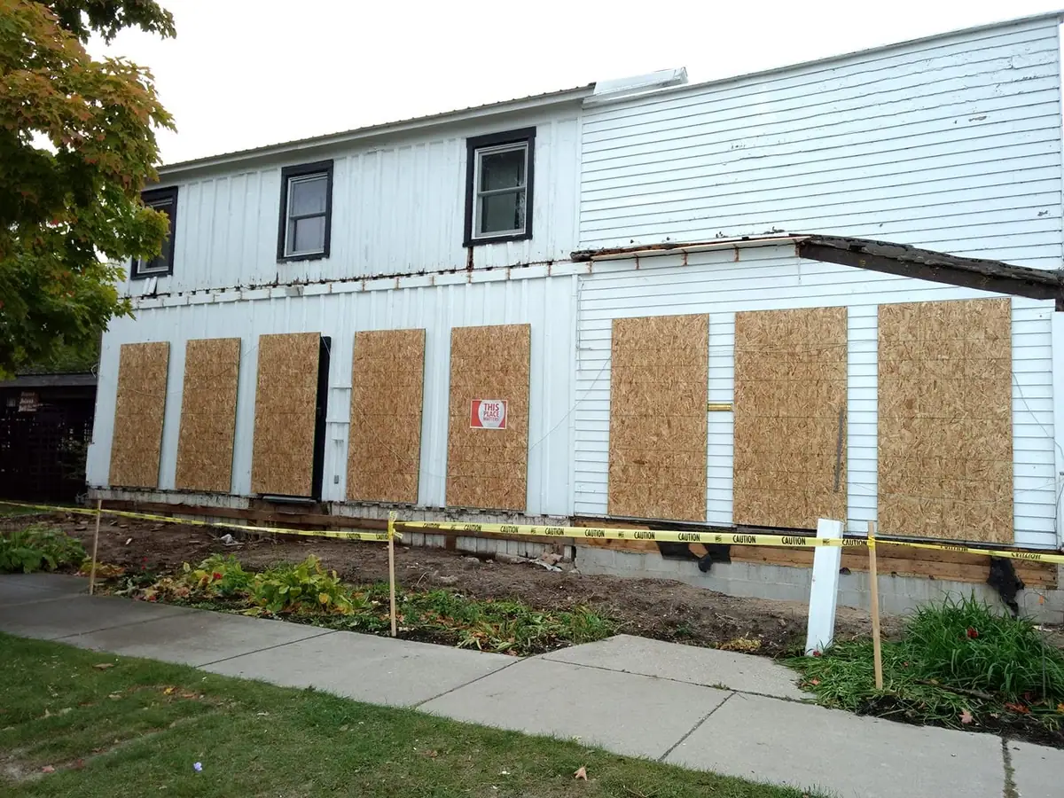 White-sided building with boarded windows, caution tape, and a “THIS PLACE MATTERS” sign.