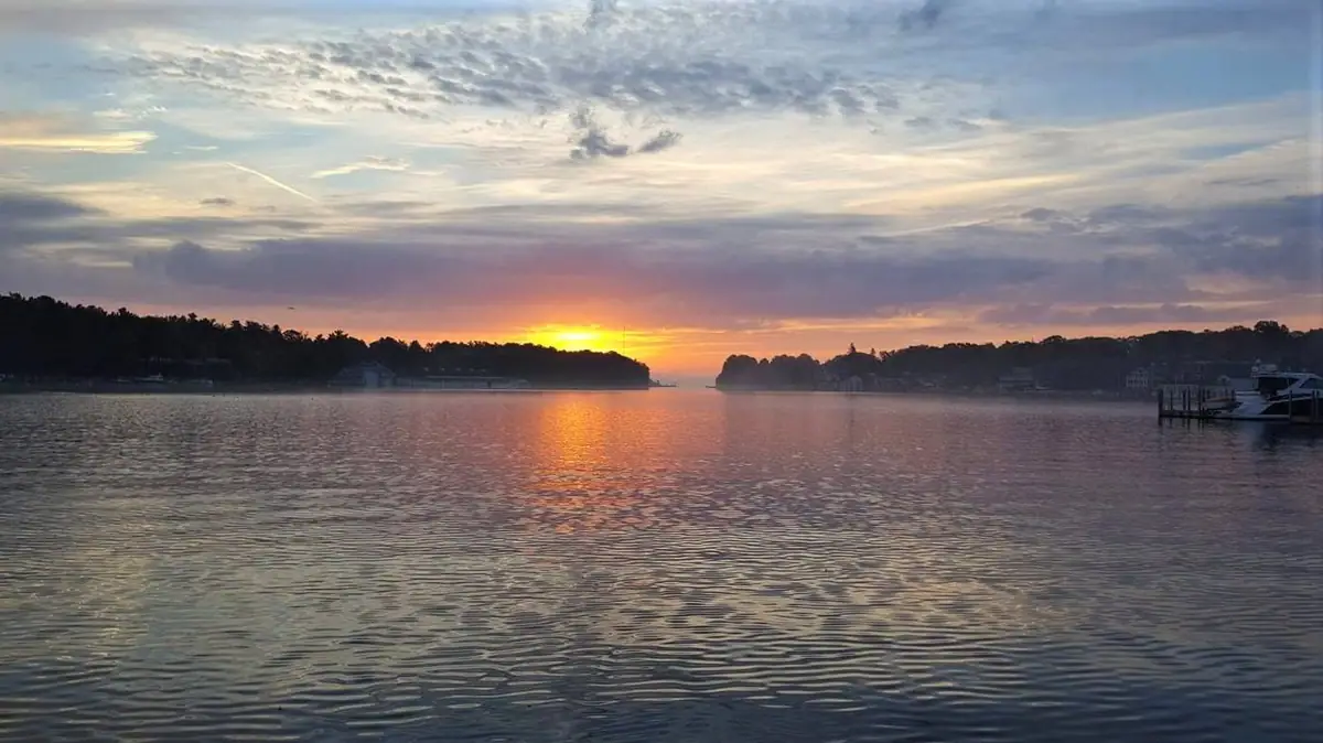 Sunrise glows over rippled water between wooded shores, with boats and docks at right.