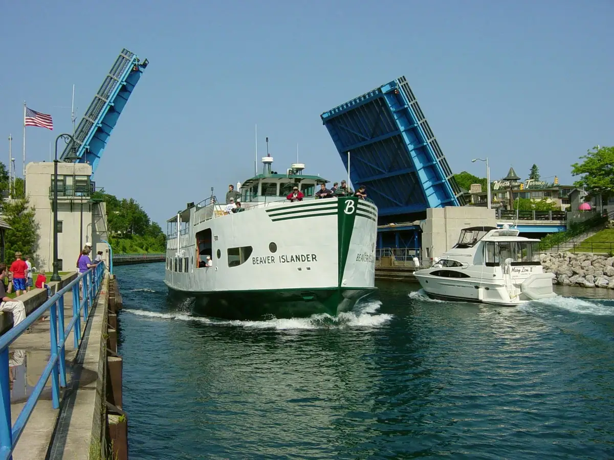 A large passenger ferry passes between the raised sections of a blue drawbridge as people watch from the pier.