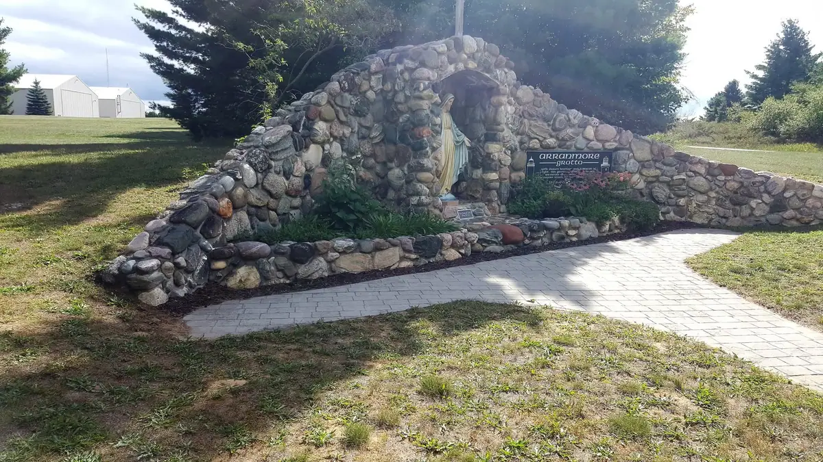 Stone grotto with a statue and sign beside a paved walkway.