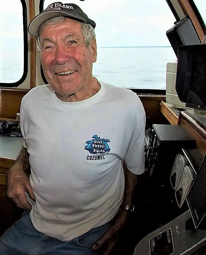 Smiling older man seated beside marine electronics inside a boat cabin, with open water visible through the windows.
