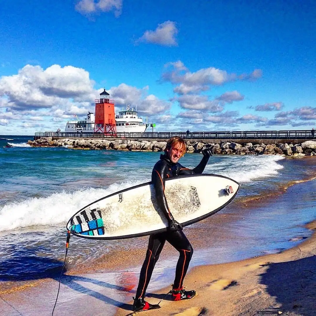 Surfer in a black wetsuit carries a white board along a sandy beach beside a pier, lighthouse, and ferry.