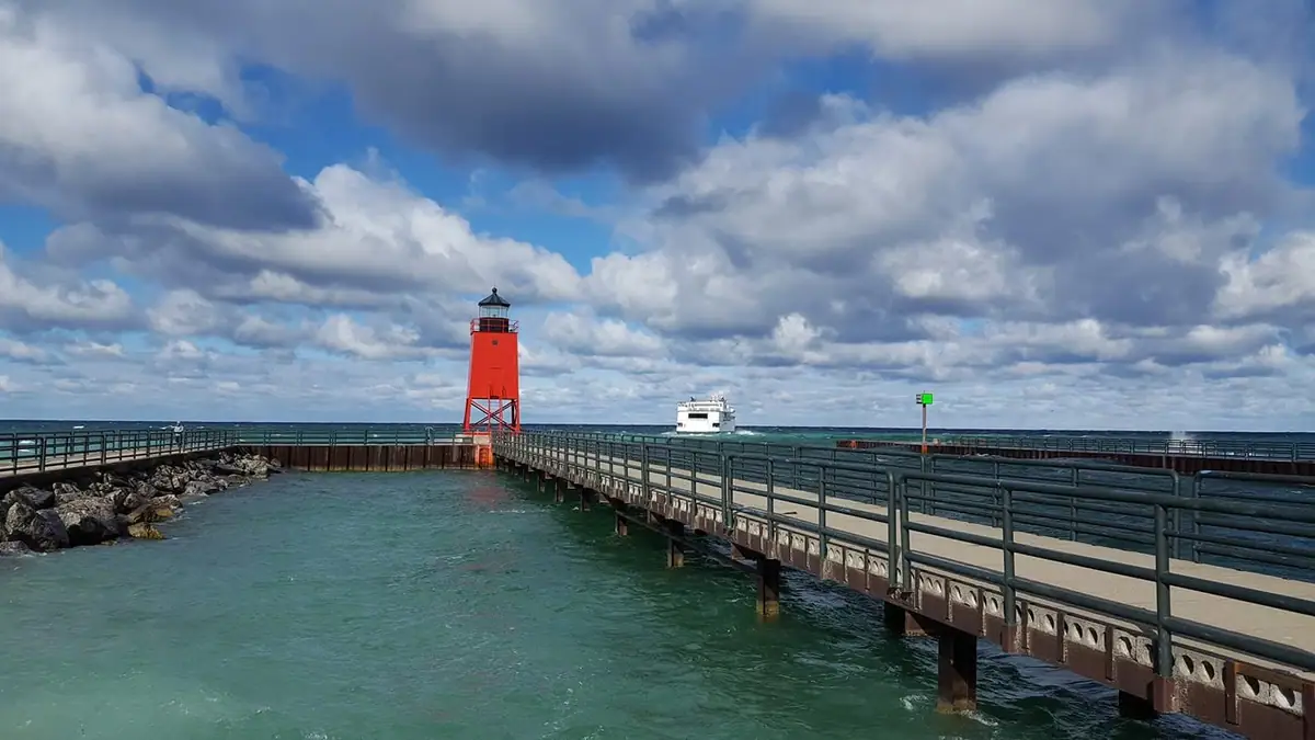 A red lighthouse stands at the end of a railed pier, with a white vessel and choppy water beneath a cloudy sky.