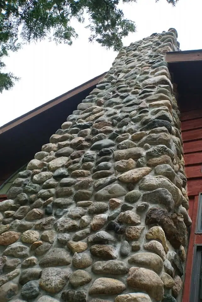 Low-angle view of a rounded fieldstone chimney rising beside a red wooden building.