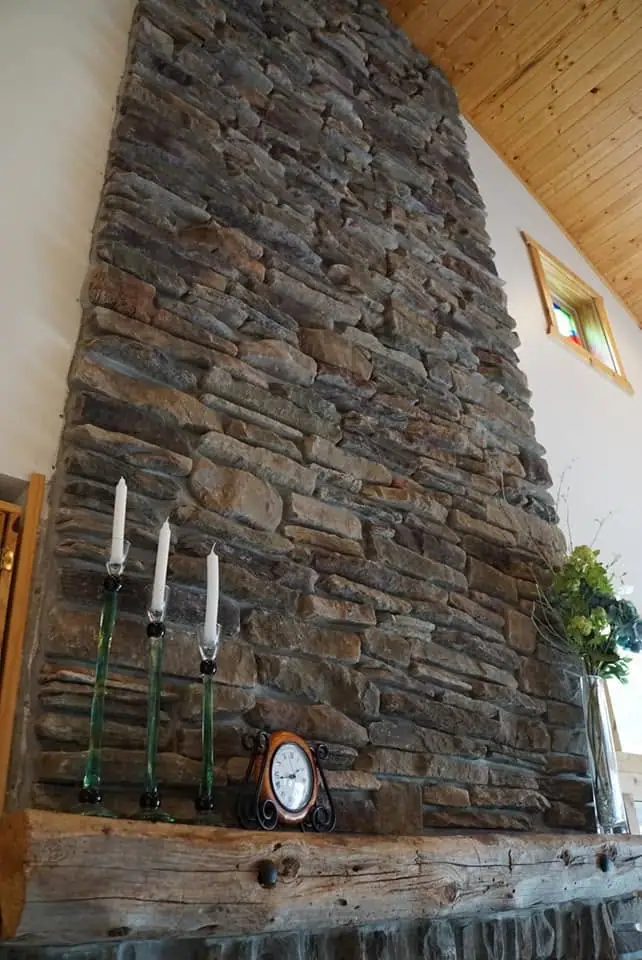 Interior view of a tall stone fireplace with a timber mantel, candles, clock, greenery, and a small stained-glass window.