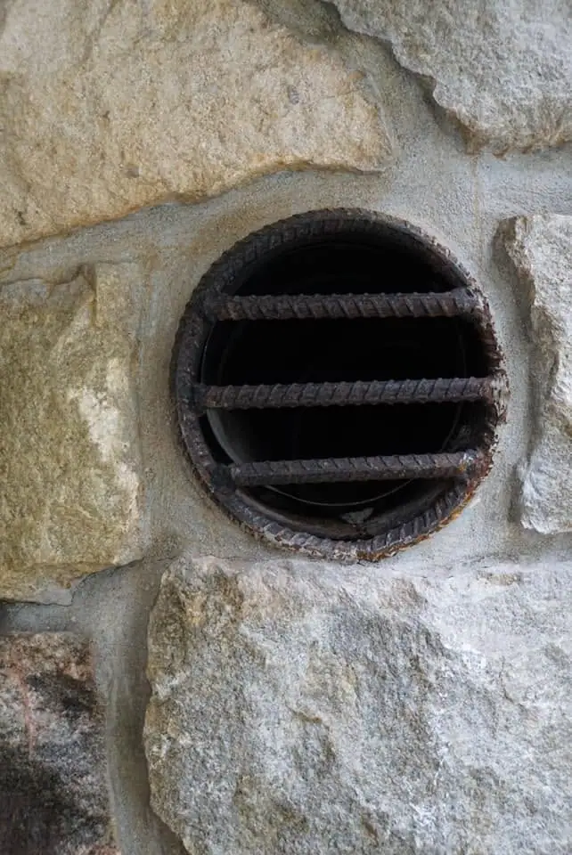 A rusted circular iron grate covers a dark opening in a rough stone wall.