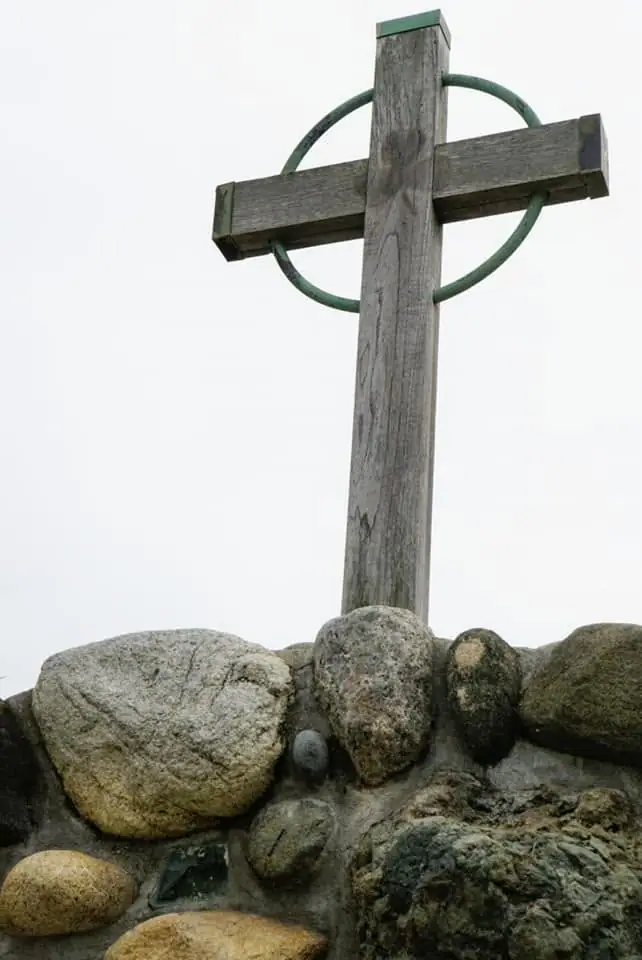 A weathered wooden cross with a green circular ring rises from a rounded-stone structure.