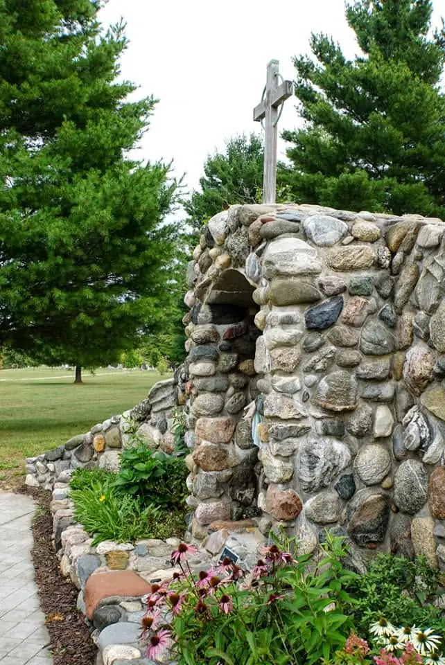 A rounded stone grotto with a wooden cross stands among flowers, evergreen trees, and a paved walkway.