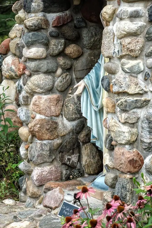 A blue-robed statue stands within a rounded-stone grotto among blooming coneflowers.