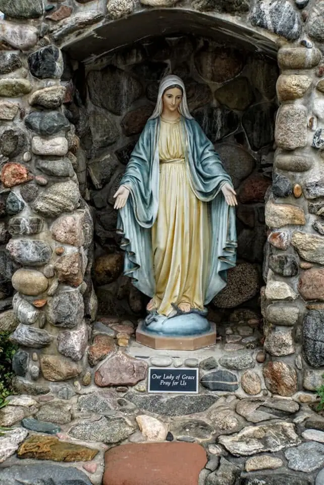Photograph of a robed female religious statue standing in a rounded stone grotto with a small plaque.