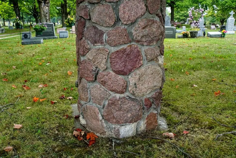 A rough stone pillar stands among gravestones and flowering plants in a grassy cemetery.