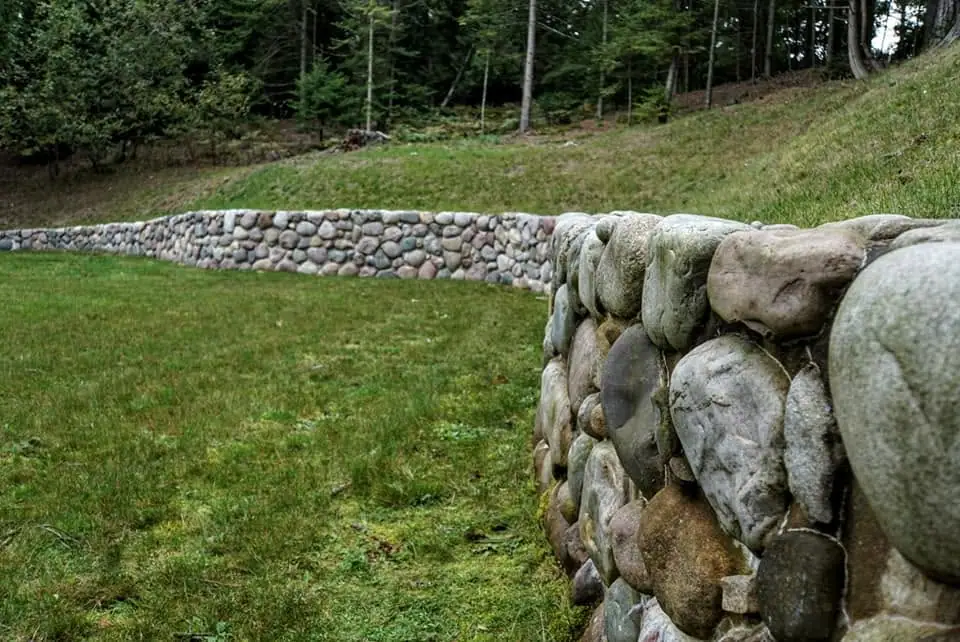 Rounded-stone retaining walls curve across a grassy slope beside a forest.