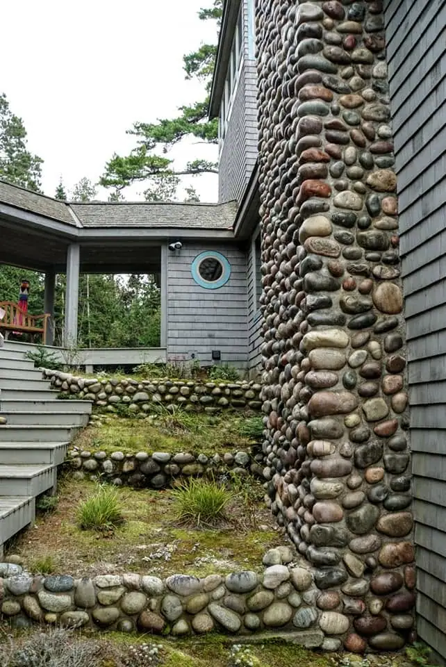 House exterior with rounded-stone walls, terraced garden beds, steps, porch, and a circular window.