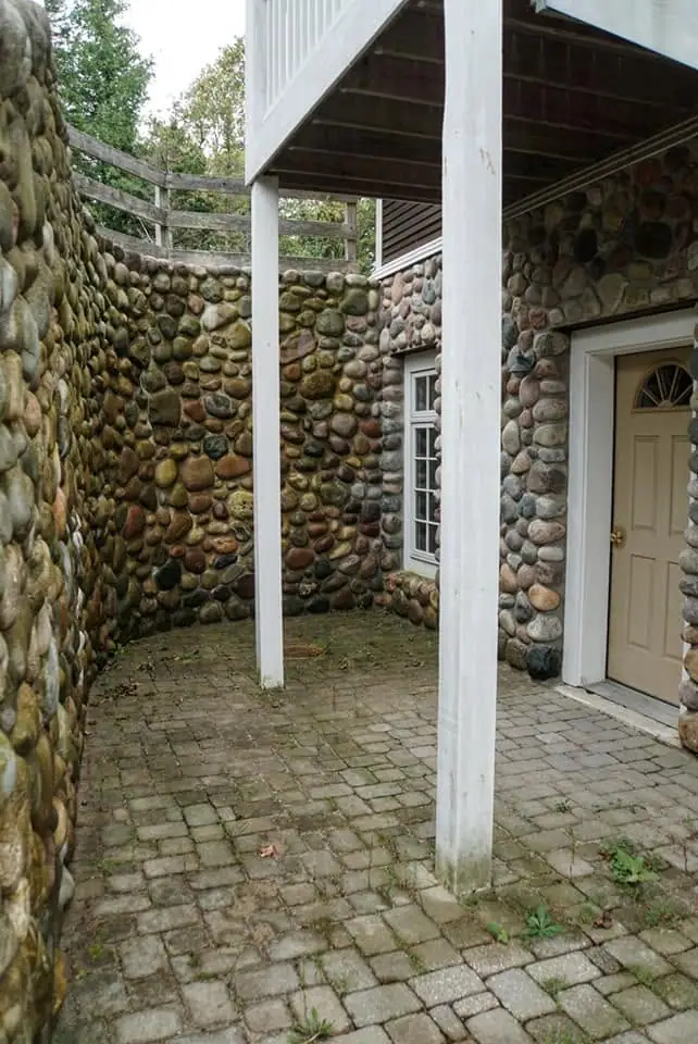 A paved entry is enclosed by rounded-stone walls, with white supports, a window, and a door.