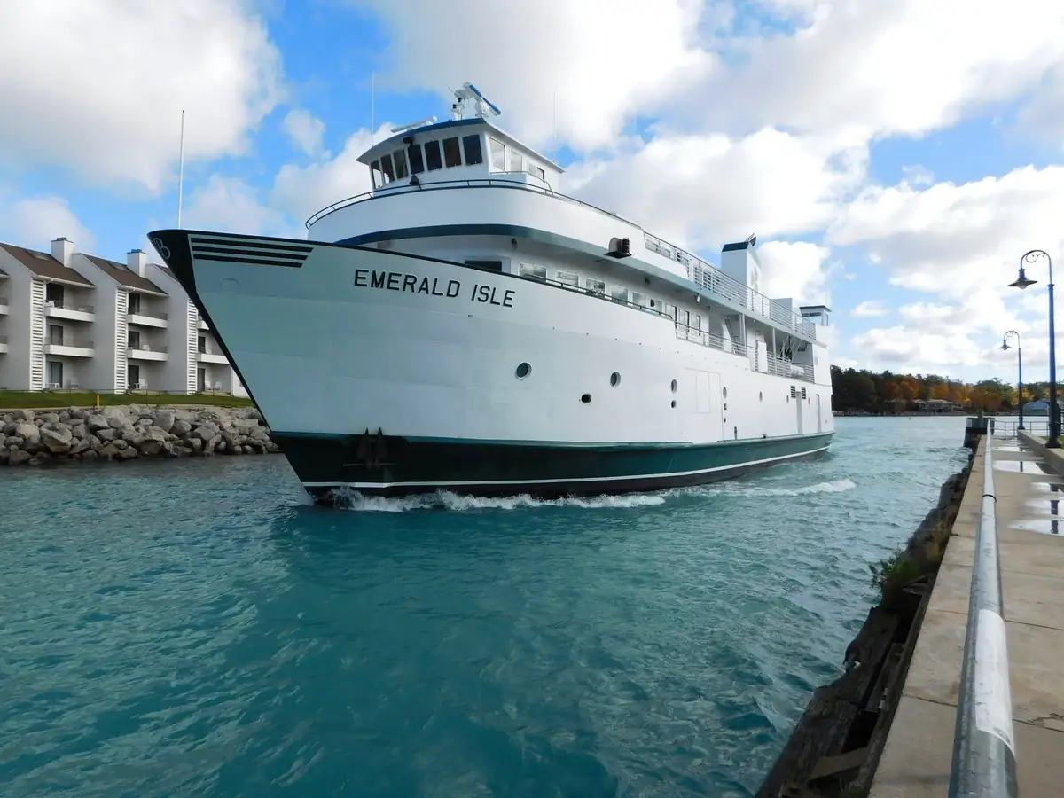 A large white ferry passes a waterfront pier beneath a bright, cloud-filled sky.
