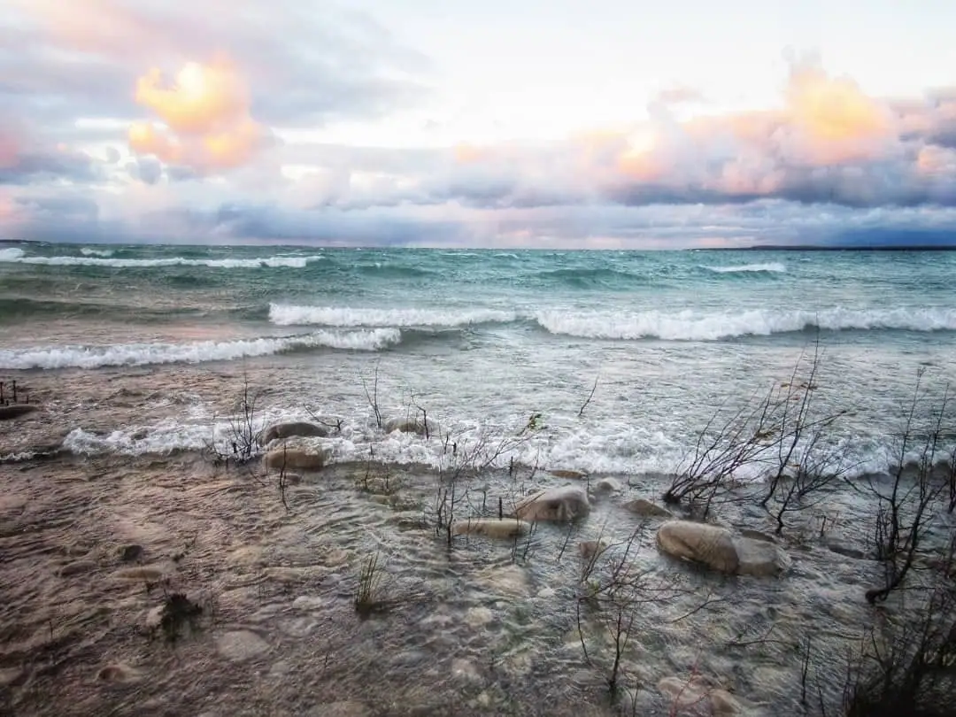 Breaking waves wash over a sandy, rocky shoreline beneath dramatic pink and gray clouds.