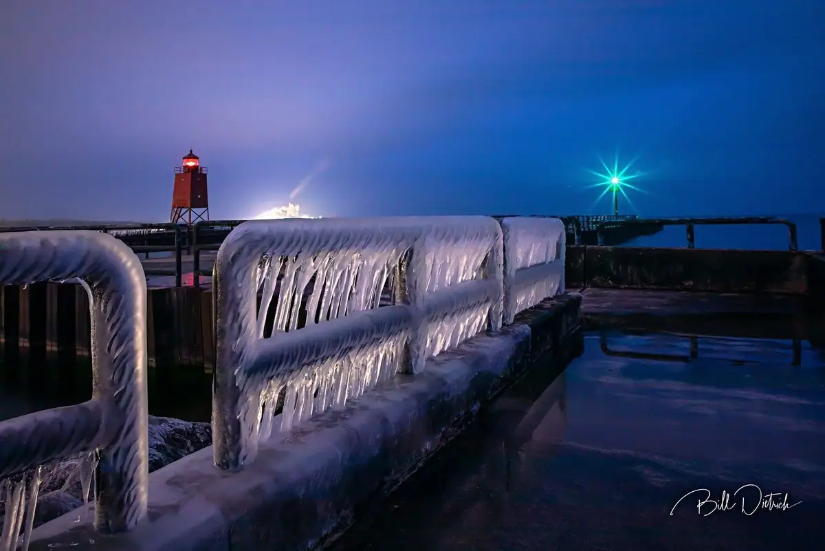Ice-coated pier railings and a lighthouse glow against the blue night sky.