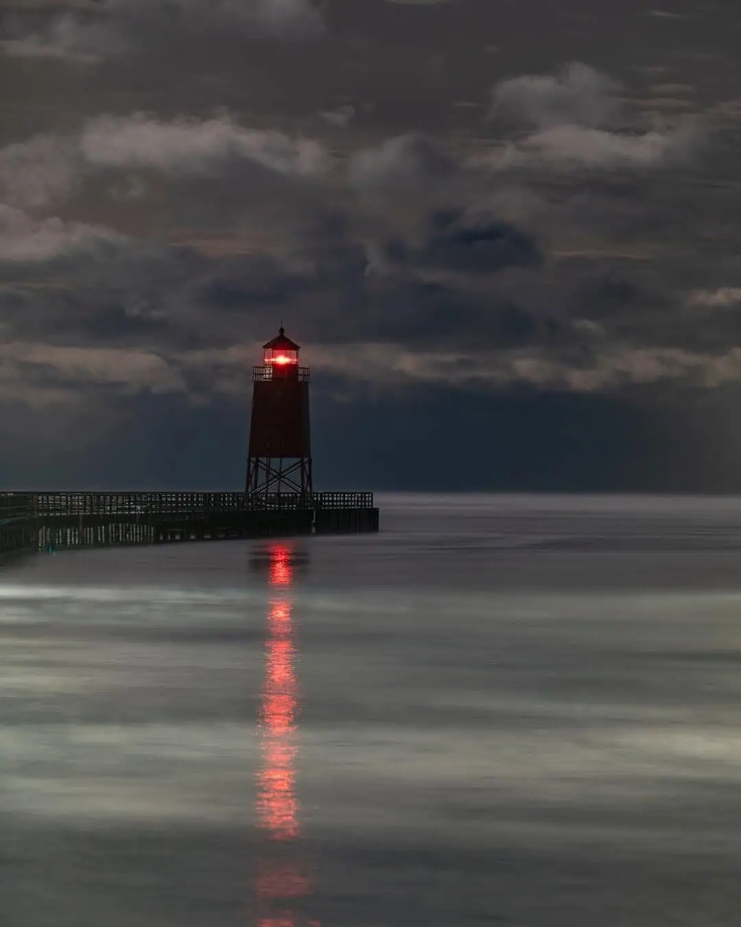 A red-lit lighthouse stands at the end of a pier, reflected in calm water beneath heavy clouds.