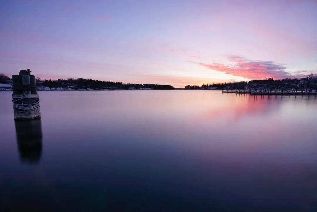 Sunset colors reflect across calm water beside a wrapped wooden post and distant shoreline docks.