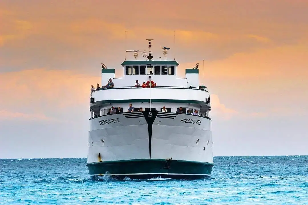 A large white passenger ferry approaches through choppy blue water beneath a warm orange-pink sky.