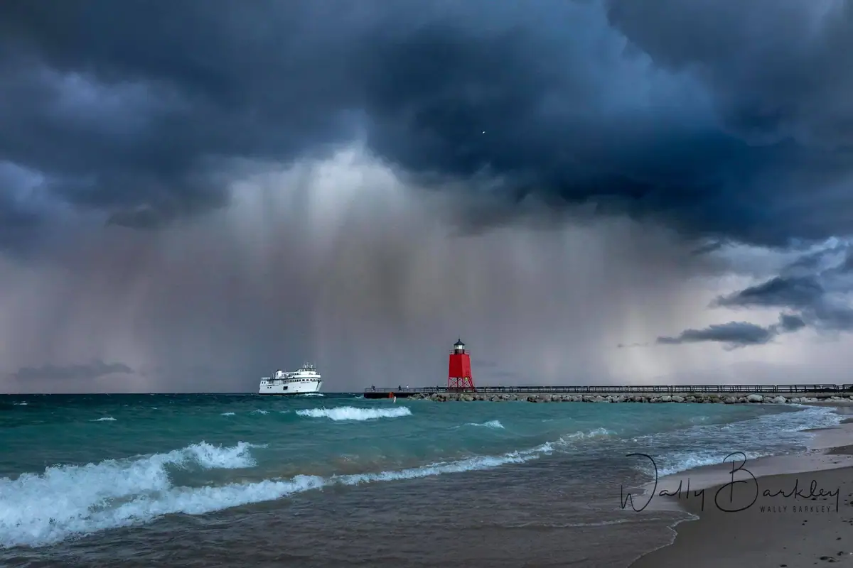 A white ferry passes a red lighthouse on a pier as waves break beneath a dark storm.