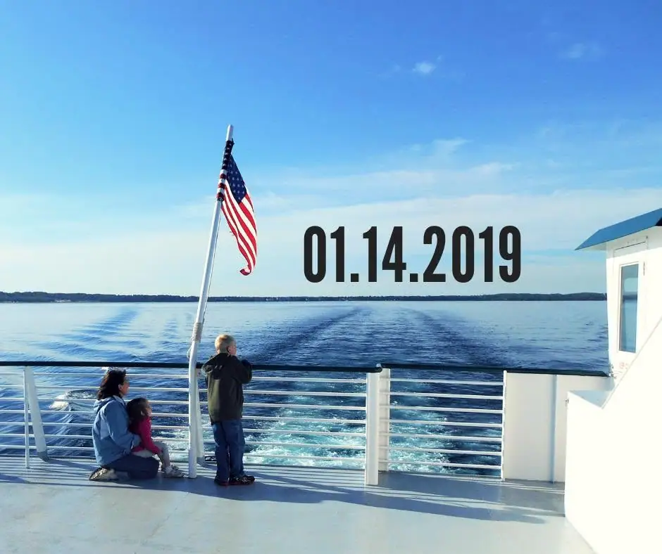 An adult and two children stand on a ferry deck beside an American flag, with wake and shoreline behind them.