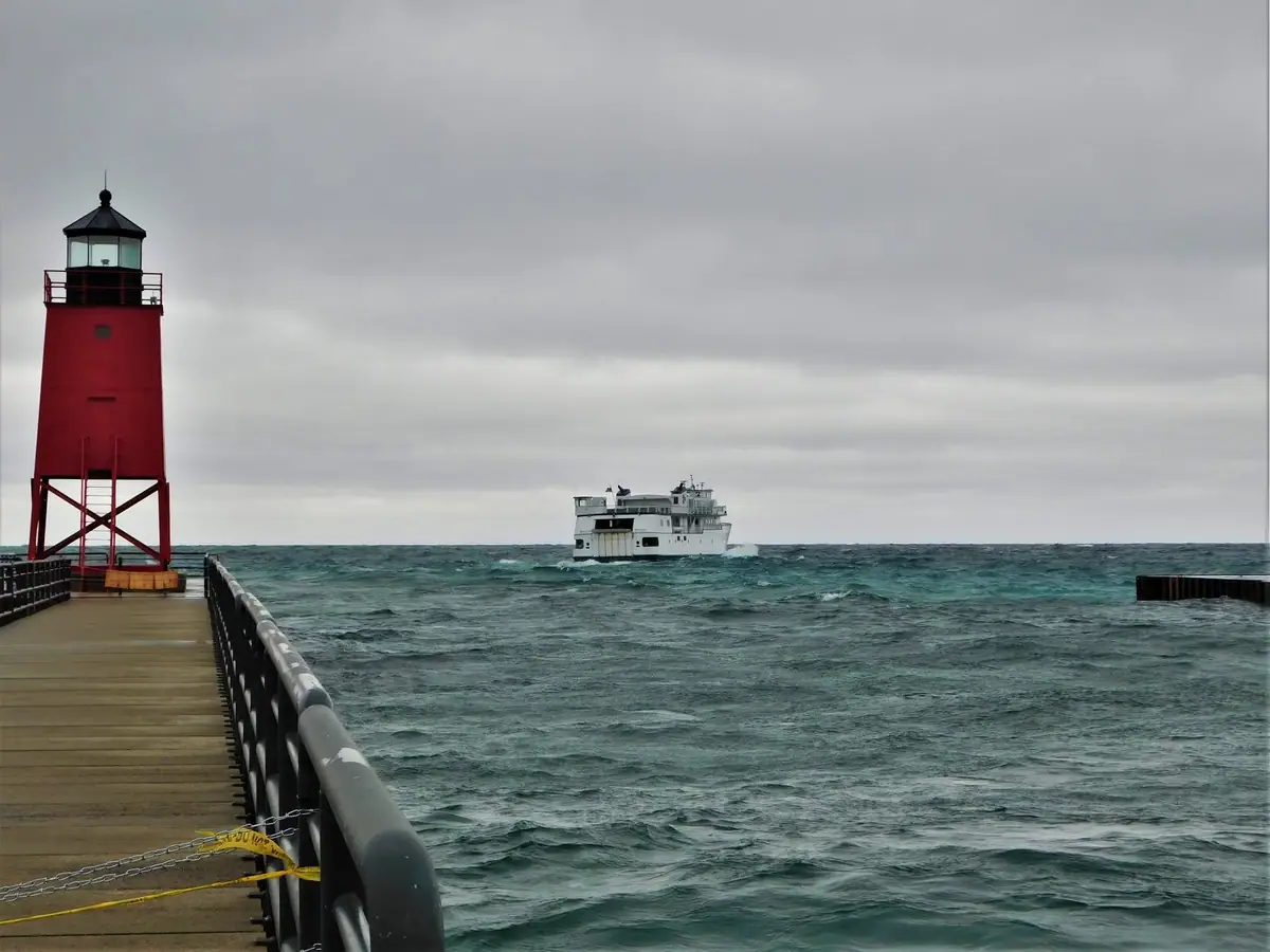 A red lighthouse stands at the end of a pier as a white ferry crosses rough water beneath gray clouds.