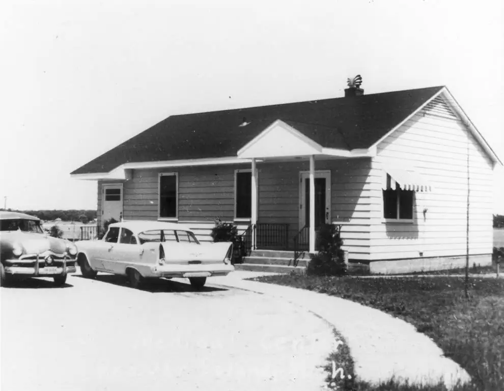 Black-and-white photograph of a single-story house with a porch and two vintage automobiles parked in front.