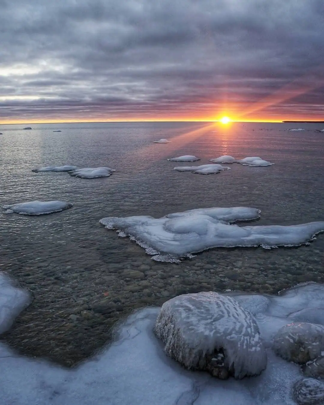 Sunset over dark water with floating ice and an ice-coated rocky shoreline.