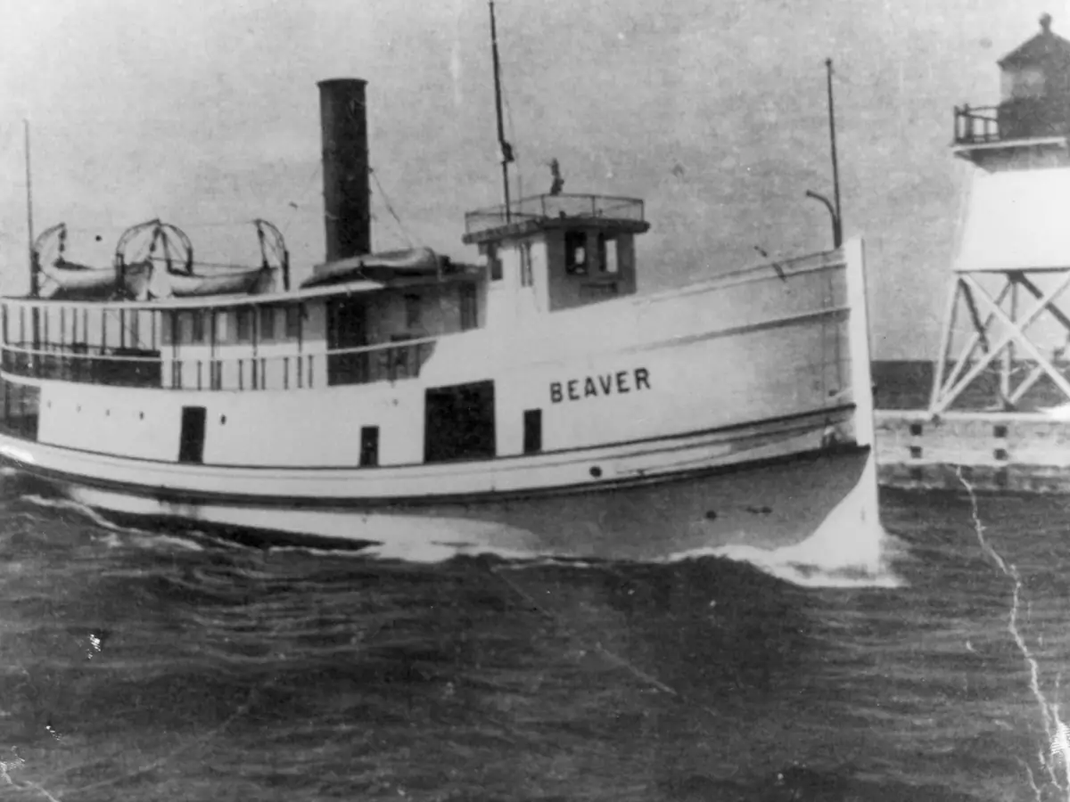 Black-and-white photograph of the ferry BEAVER moving through choppy water beside a lighthouse.