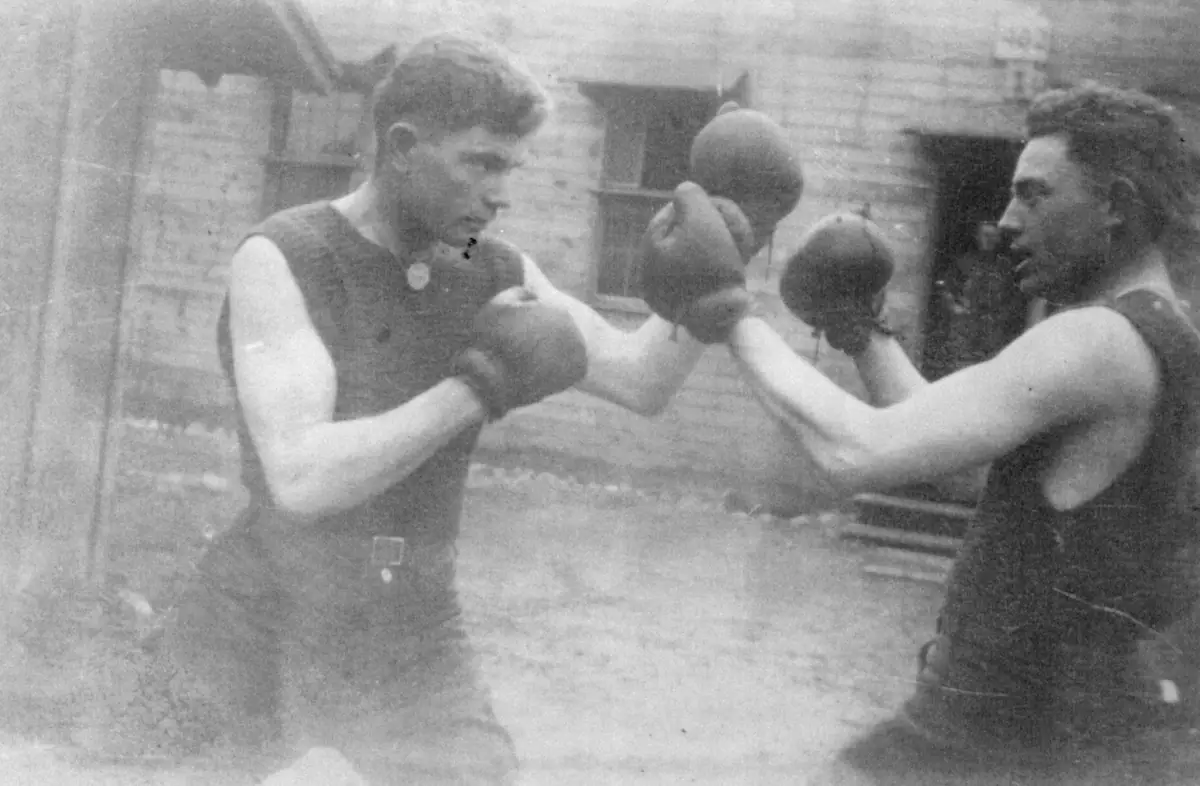 Two men wearing boxing gloves spar outdoors in front of a wooden building.