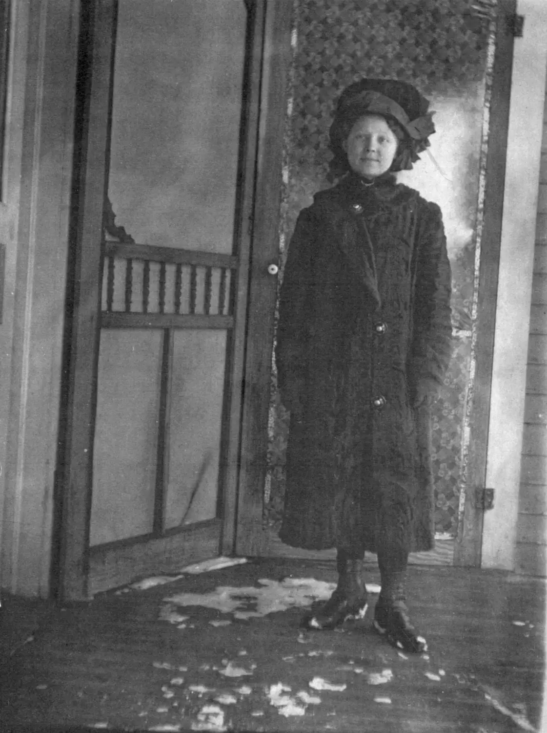Black-and-white portrait of a woman in a dark coat and large bow-trimmed hat standing in a doorway.