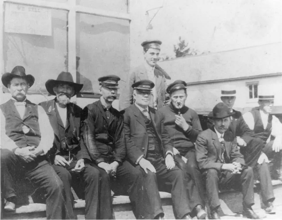 Nine men in hats and suits pose outside a building, with faint lettering visible on an upper-left sign.