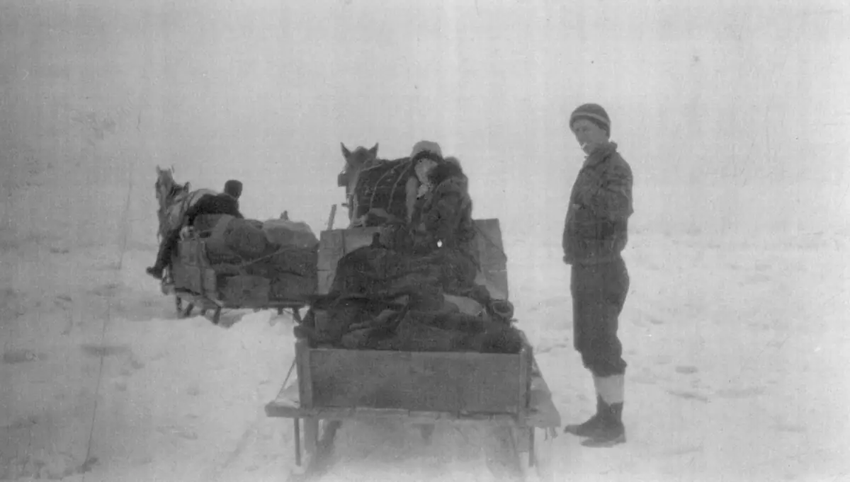 Two horse-drawn sleds carrying bundled loads stand in a snowy open landscape with several people.